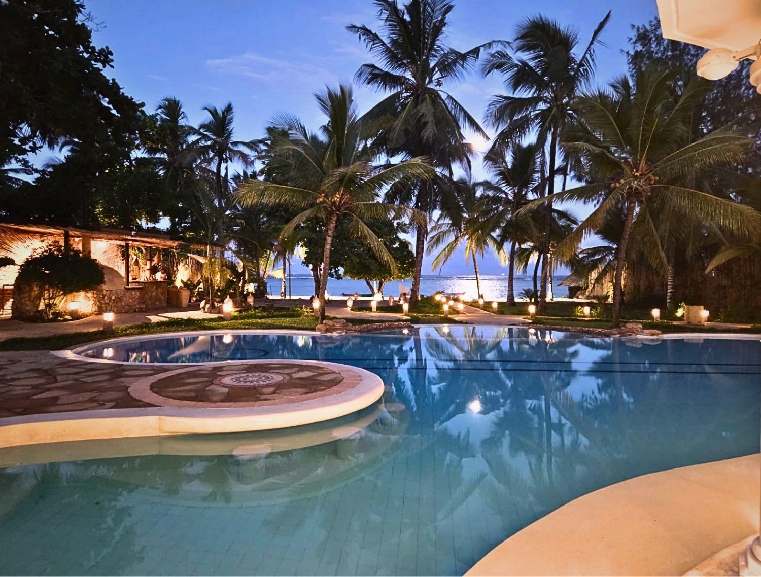 Poolside at a tropical resort at dusk, with palm trees, warm lights, and the ocean visible in the background. - Soroi Collection