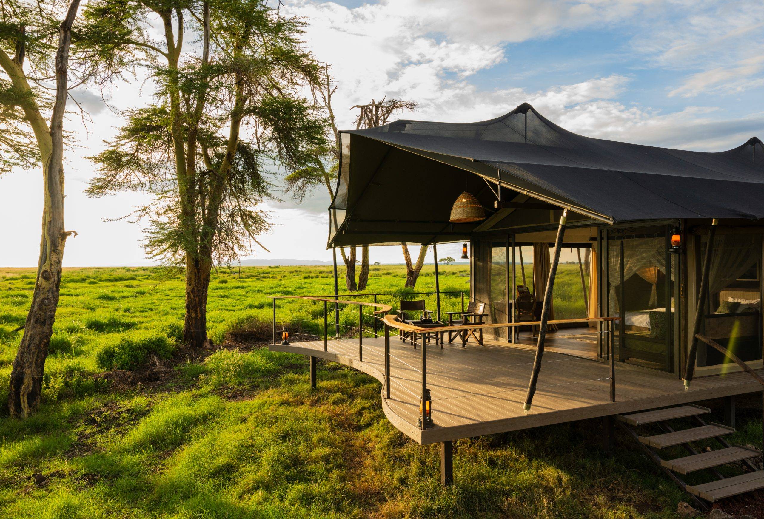 Tented lodge on a wooden deck in a grassy savanna, with trees in the background and outdoor seating under a dark canopy. - Soroi Collection - Soroi Collection