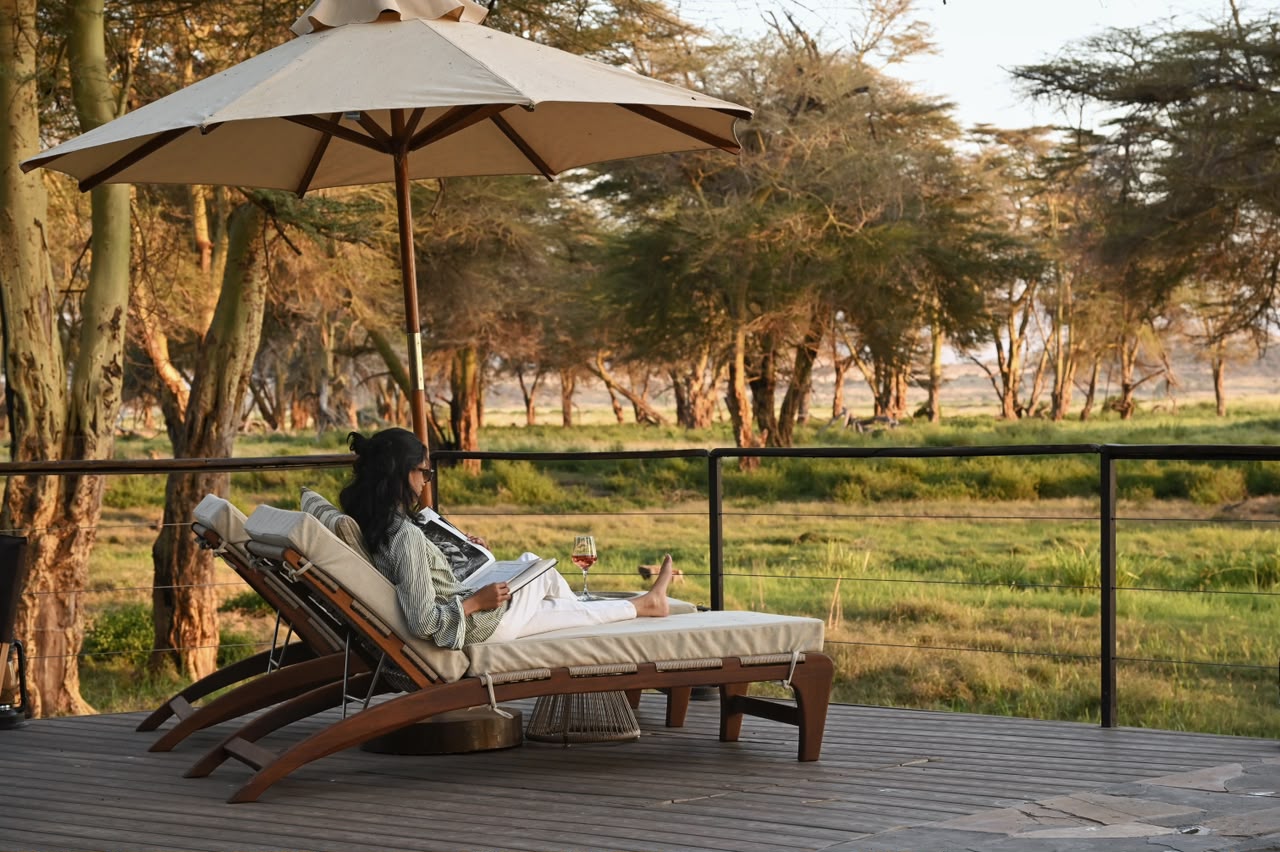 Person lounging on a cushioned outdoor lounge chair under a large umbrella on a wooden deck, reading a magazine with a glass of wine nearby. - Soroi Collection