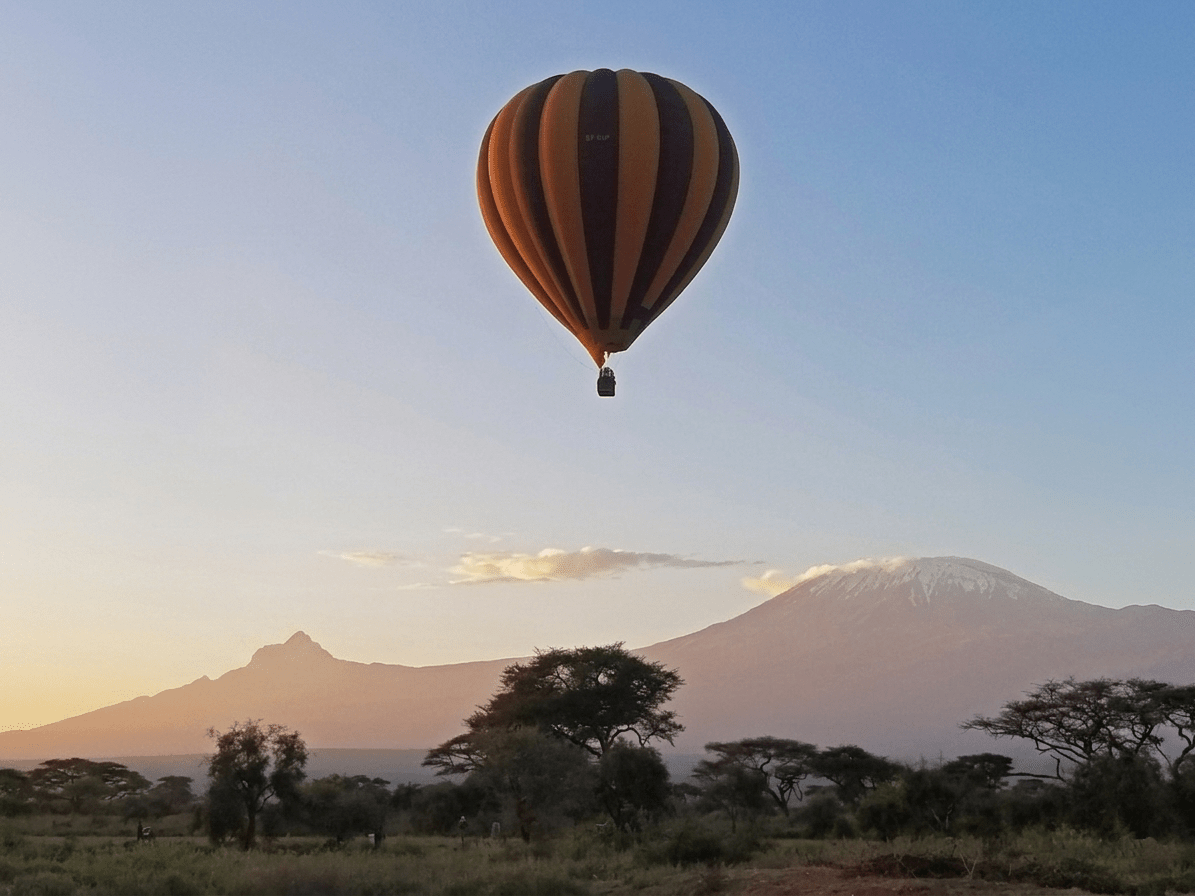 Striped orange and black hot air balloon drifting over a savannah with distant snow-capped mountain at sunset. - Soroi Collection - Soroi Collection