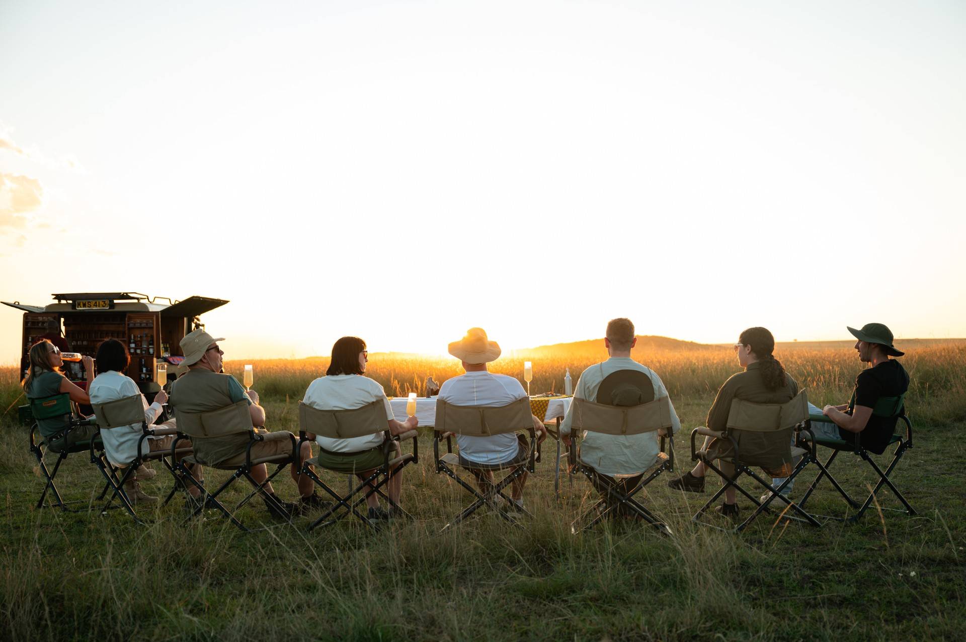 Group of people sitting in folding chairs around a long table in a grassy field at sunset, drinks and candles on the table. - Soroi Collection - Soroi Collection